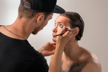 Şapkalı MUA ressamı, stüdyosunda göğsünde dövme olan sakallı genç bir aktöre göz kalemi uyguluyor.