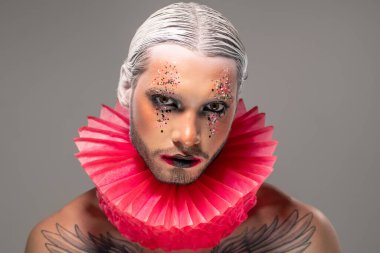 Beyaz saçlı, siyah dudaklı, gri arka planda kameraya bakan depresif genç sakallı aktörün portresi.