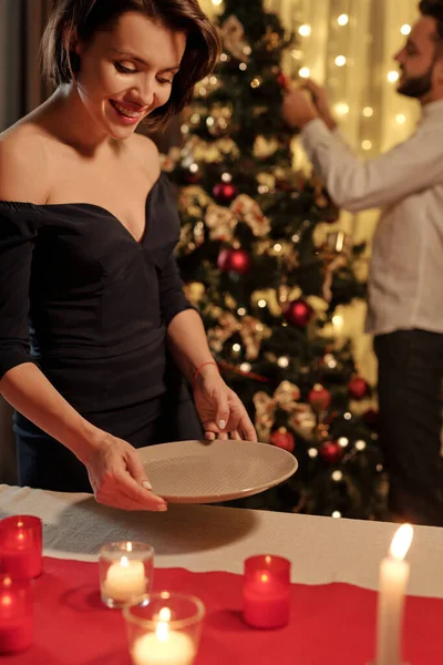 笑顔励起若いです女性で黒ドレスサービングクリスマステーブルのために夕食中に彼女のボーイフレンド装飾木で背景 — ストック写真