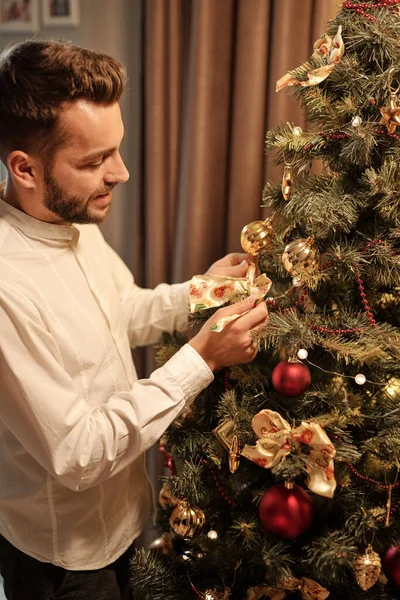 白いシャツを着た若い男がクリスマスツリーに立って リビングルームで木の上でクリスマスの装飾を調整する — ストック写真