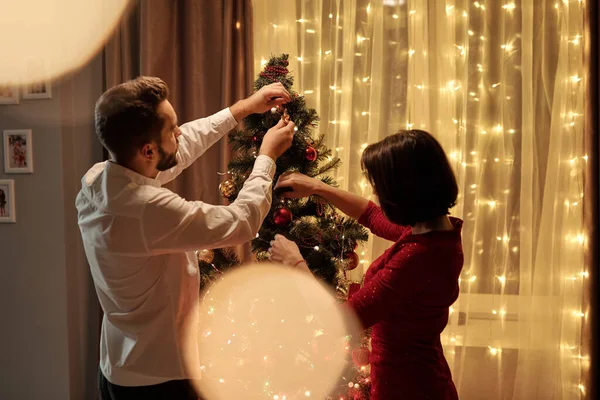 クリスマスツリーに立っている若いカップルの背面ビューと新年の準備中におもちゃをぶら下げ ライト効果 — ストック写真