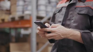İnşaat elbisesi giymiş işçinin, depoda yürürken akıllı telefon mesajları tutarken tanınmayan ellerinin orta bölüm yavaş çekim görüntüsü.