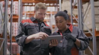 Kafkas inşaat işçisi Afrika kökenli Amerikalı kadın işçilere iş planını açıklarken orta boy bir fotoğraf hem tulum giyerken hem de büyük bir depoda duran tablete bakarken.