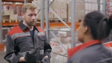 Erkek müteahhitin orta boy yavaş çekimi. Depoda kutularla dolu raflarla çalışan Afrikalı Amerikalı bir kadınla konuşuyor.