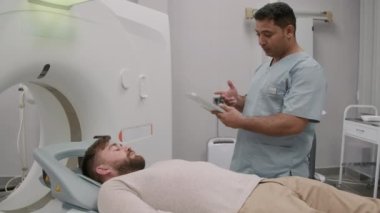 Önlüklü Arap erkek teknolojisinin yavaş takibi tablet kullanıyor ve CT tarama makinesinde kanepede yatan hastayla konuşuyor.