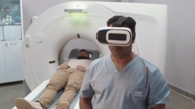 Önlüklü, VR kulaklık takan ve radyoloji odasında görünmez arayüz kullanan Arap erkek teknolojisinin orta boy fotoğrafı. Hasta CT taramasında tedavi koltuğunda yatıyor.