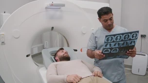胸部X線を保持し Ctスキャンで治療ソファの上に横たわる患者と話すアラブの男性放射線技師のハンドヘルド追跡 — ストック動画