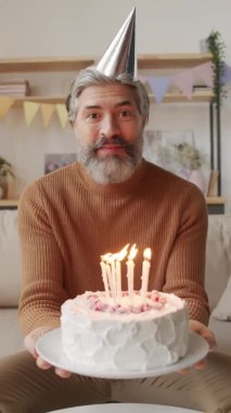 Beyaz tenli orta yaşlı bir adamın doğum gününü videolu sohbet ile kutlarken doğum günü pastasında mumları üflerken gülümsediği dikey bir görüntü.