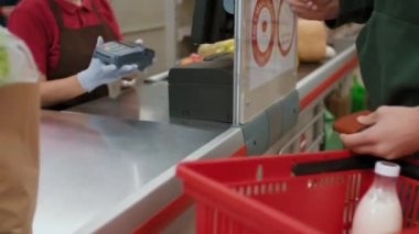 Kırmızı üniformalı, koruyucu eldivenli, tanınmayan bir kadın kasiyerin, müşteriye ulaşamadığı bir şekilde kredi kartıyla ödeme yaptığı orta bölüm görüntüleri.