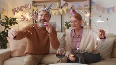 Orta ölçekli mutlu orta yaşlı maytap ve şenlikli partileri olan orta yaşlı bir çiftin görüntüsü. Evdeki dekore edilmiş oturma odasında video aracılığıyla kutlama yapıyorlar.