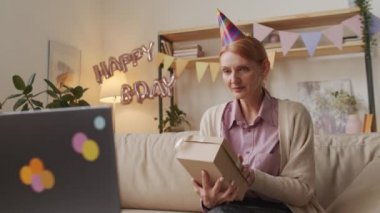 Orta yaşlı bir kadının doğum gününü online kutlayan video konuşmasını gösteren orta ölçekli yavaş çekim görüntüleri. Evde oturmuş, hediye kutularını açarken dizüstü bilgisayara bakarken.
