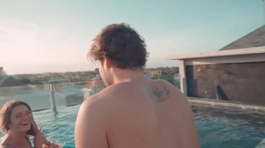 Keyifli bir adam havuzun kenarında oturuyor ve kız arkadaşı güneşli bir günde lüks bir tatil köyünde eğleniyor.