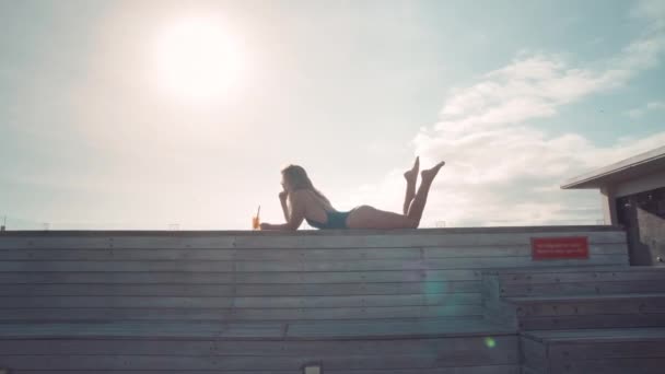 穿着绿松石泳衣的美女躺在肚子里 摆动着腿 喝着鸡尾酒 望着蓝天 — 图库视频影像