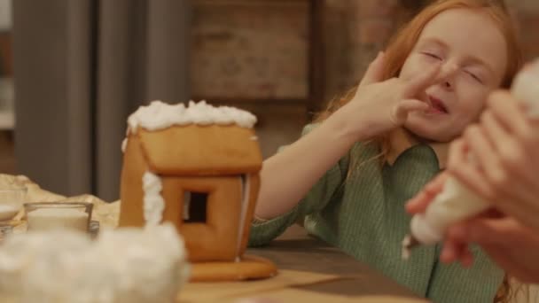 テーブルに座っている素敵な女の子のスローモーションのクローズアップ自家製のジンジャーブレッドハウスの試飲バタークリームの笑顔とクリスマスを楽しんで — ストック動画