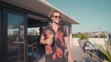 Orta ölçekli bir fotoğraf, elinde kokteylle kız arkadaşına yaklaşan ve kamerayla konuşan bir adam görüntüsü.