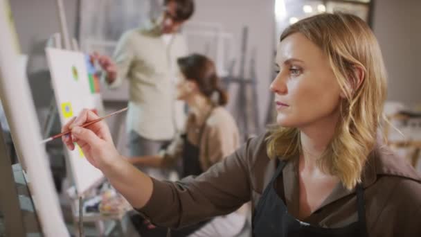在明亮的现代艺术工作室里 男性艺术导师在与学生进行背景对话时 用画笔在画布上进行集中的漂亮女子绘画的中等特写 — 图库视频影像