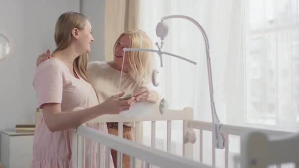 中慢镜头拍摄的快乐的年轻孕妇 与未来的祖母站在婴儿床边装饰着自制婴儿床玩具笑着和说话 — 图库视频影像