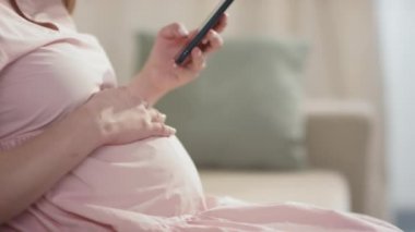 Karnına dokunan ve akıllı telefon kullanan tanınmayan hamile kadının orta bölüm yavaş çekim yan görüntüsü.