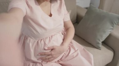 Hamileliği hakkındaki haberleri paylaşırken gülümseyen mutlu, genç bir kadının el kamerasıyla büyük göbeğini gösteriyor.