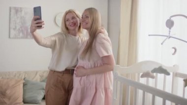 Orta ölçekli, neşeli, iyi görünümlü, hamile kızıyla özçekim yapan, arka planda beyaz bebek yatağı ve beşik oyuncakları olan bir büyükannenin fotoğrafı.