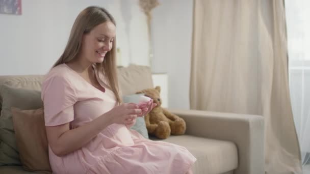 中等慢镜头 一个微笑的孕妇坐在客厅的沙发上 抱着小女婴的靴子看着她们 — 图库视频影像