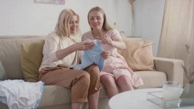 Genç hamile bir kadının, mutlu bir anneye yeni doğmuş kıyafetlerini gösterirken, oturma odasındaki kanepede yan yana otururken orta yavaş çekilen görüntüler.