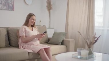 Kafkasyalı hamile bir kadının pastel renkli oturma odasındaki koltukta tek başına oturmuş akıllı telefonuna bakarken gülümsediği orta ölçekli yavaş çekim görüntüleri.