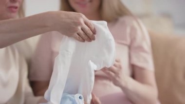 Oturma odasındaki kanepede oturan ve ellerinde yeni doğmuş bebek kıyafetleriyle tanınmayan iki kadının orta bölüm yavaş çekim görüntüleri.