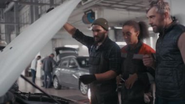 Orta yaşlı bir adam sorunu anlatırken, erkek oto tamircisi ve bayan arkadaşının araba kaputunun altında konuşurken ve bakarken elde çekilmiş bir fotoğrafı.