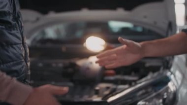 Tanımlanamayan erkek müşteri ve erkek oto tamircinin yavaş yavaş el sıkışması. Kaputu açık ve arka planda taşınabilir iş lambası olan bozuk bir araba.