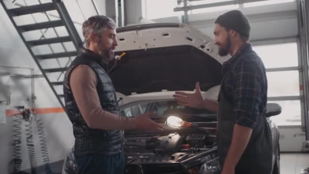 身着工作服的男性汽车修理工和留着胡子的男性顾客站在车前 打开车篷并在车库里握手的慢镜头 — 图库视频影像