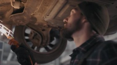 El kamerasıyla çekilen düşük açılı sakallı erkek araba tamircisi portatif iş lambası kullanıyor ve arabanın bagajında tamirat yapıyor.