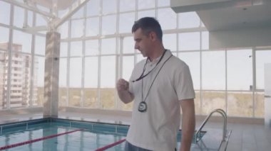 Beyaz polo tişörtlü, havuzda yüzme antrenmanı yapan ve ıslık çalan orta yaşlı bir adam.