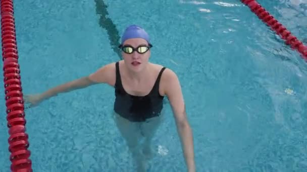 身穿泳衣 头罩和护目镜的年轻女运动员站在游泳池的水里 看着镜头的高角慢镜头 — 图库视频影像