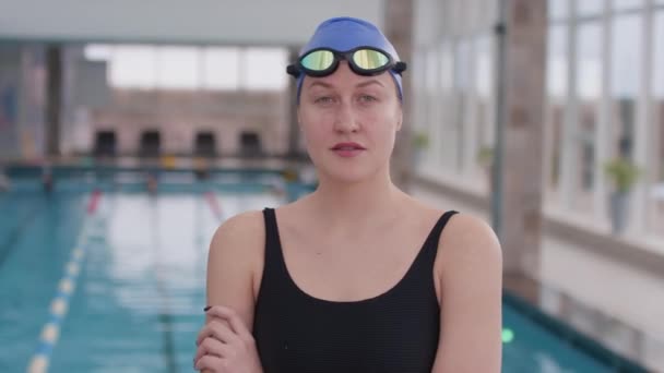 戴着帽子和护目镜在室内游泳池看着相机的年轻女子游泳运动员的慢镜头肖像画 — 图库视频影像