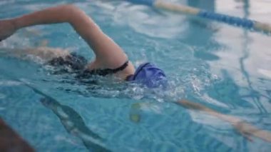 Mayo, kep ve gözlüklü kadının havuzda kurbağalama darbeleriyle yüzüşünün yavaş takibi.