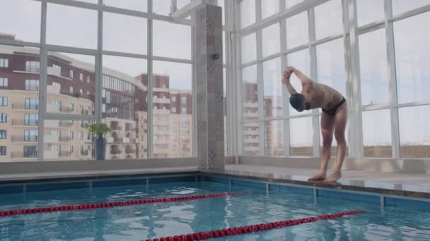 一排排的射击镜头 男子泳衣 护目镜和泳裤慢吞吞地伸展和热身 然后跳进室内游泳池 — 图库视频影像