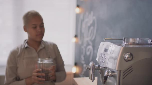 在室内小咖啡屋工作的年轻快乐黑人妇女将新鲜咖啡豆倒入电动咖啡机 准备开咖啡店的侧面镜头 — 图库视频影像