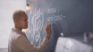 Siyah tahtada tebeşirle kahve, cappuccino, latte yazan, küçük bir kafede çalışan Afrikalı Amerikalı genç bir kadının orta boy yan görüntüsü.