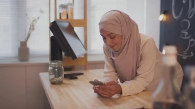 Gülümseyen müslüman kadının iş yerinde akıllı telefon kullanarak kasiyer masasına eğildiği ve küçük kapalı kafeteryalarda kahve sattığı bir fotoğraf.