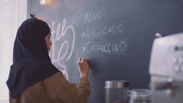 顧客のための黒板にコーヒー飲料を書いてヒジャーブの若いイスラム教徒のコーヒーショップの労働者の中期側のビューのクローズアップ — ストック動画