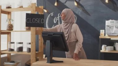 tesettürlü neşeli genç müslüman kadının orta boy portresi küçük kafe açık tabela panosunda işyerine varıyor kameraya bakıyor ve müşterilerin gülümsemesini bekliyor.