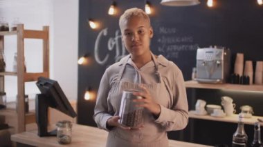 Bej önlüklü Afro-Amerikan kahve dükkanı çalışanının orta boy portresi. Bir kavanoz kahve çekirdeği ile kameraya bakıp gülümsüyor.