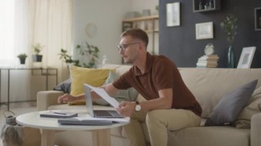 Gözlüklü, ciddi bir beyaz adamın kanepede oturmuş evrak işleriyle uğraşırken ve oturma odasında dizüstü bilgisayarla çalışırken orta boy fotoğrafı.