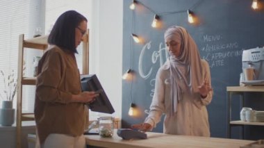 Orta boy Asyalı bir kadının küçük bir kafeden kahve alırken akıllı telefondaki temassız ödemeyi kullanarak dost canlısı Müslüman kahve satıcısına gülümserken görüntüsü.