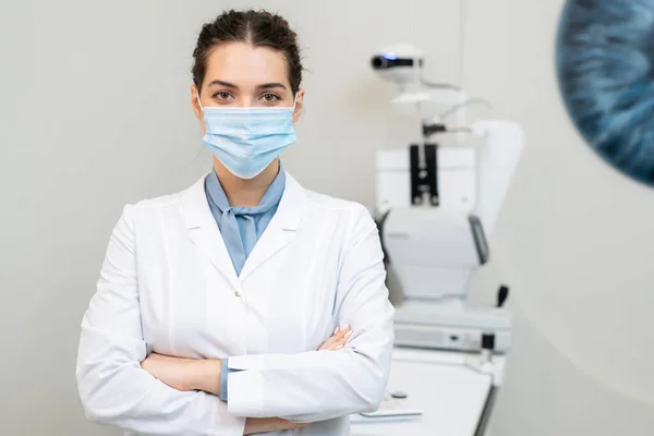 年轻的黑发女眼科医生戴着防护面罩 站在医疗器械旁边 双手叉腰穿刺 — 图库照片