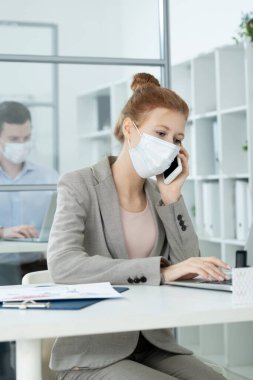 Koruyucu maskeli genç ve güzel bir iş kadını bilgisayarına bakıyor. Verilerle çalışıyor ve meslektaşına karşı ofisini arıyor.