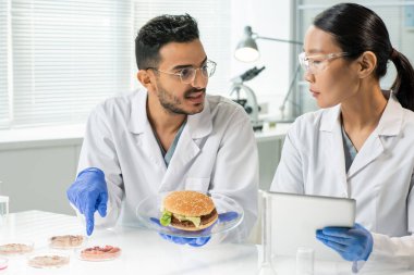 Eldivenli genç adam, tabağında hamburgerle çiğ sebze eti örneklerinden birini gösterirken meslektaşına karakteristik özelliklerini anlatıyor.