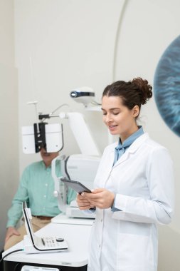 Whitecoat 'taki mutlu genç bayan göz doktoru yaşlı erkek hastanın arka planındaki tablet görüntüye bakıyor.