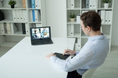 Genç zarif iş adamı ofis ortamında masanın başında oturuyor ve iş arkadaşlarıyla çevrimiçi iletişim kurarken dizüstü bilgisayara bakıyor.